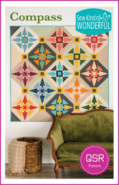 May include: A quilt pattern called 'Compass' by Sew Kind of WONDERFUL. The quilt is made of colorful fabric squares in shades of orange, yellow, blue, green, and pink. The pattern is a geometric design with a compass-like shape in the center of each square. The quilt is displayed on a white wall with a green velvet sofa and a wicker basket with a tan blanket in the foreground. The text 'QSR Pattern' is in a pink circle with white polka dots.