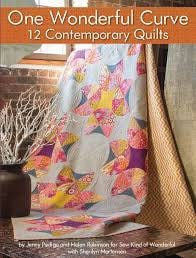 May include: A quilt with a contemporary design featuring a large, curved, gray and white circle surrounded by smaller, colorful circles and stars. The quilt is made with a variety of fabrics in shades of pink, purple, yellow, and orange. The quilt is displayed on a brick wall with a vase of white branches in the foreground. The book title 'One Wonderful Curve' and '12 Contemporary Quilts' are visible at the top of the image.