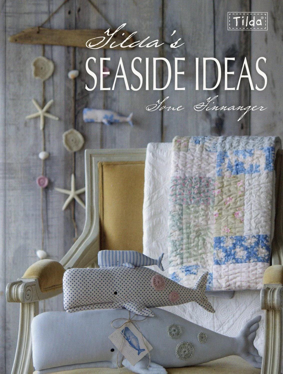 May include: A decorative display featuring a book titled "Gilda's Seaside Ideas" and whale-shaped plush toys. The toys are in various sizes and patterns, including blue stripes and white polka dots. A quilt with blue and white floral patterns is draped over a chair.