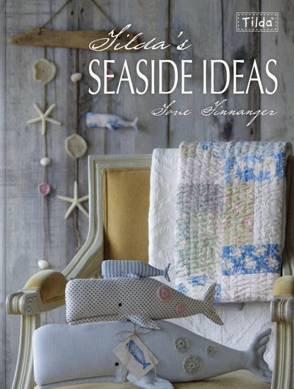 May include: A decorative display featuring a book titled "Gilda's Seaside Ideas" and whale-shaped plush toys. The toys are in various sizes and patterns, including blue stripes and white polka dots. A quilt with blue and white floral patterns is draped over a chair.